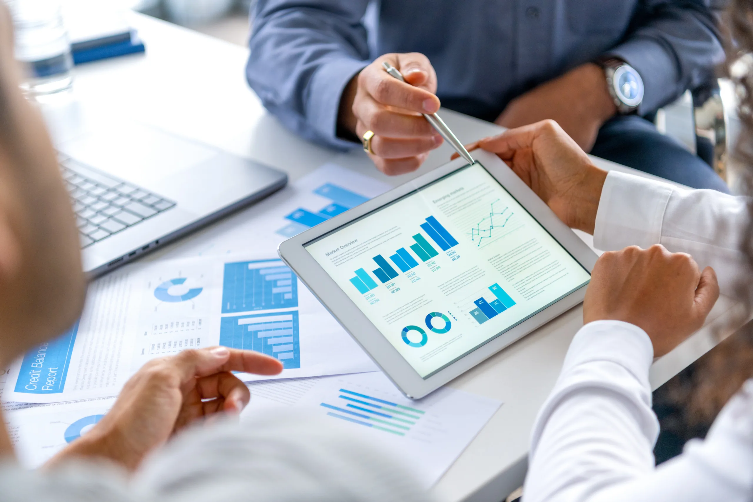 Close up of three people looking at financial data with graphs and charts. All their hands can be seen and one person is pointing with a pen. There is paperwork on the desk showing more finance information