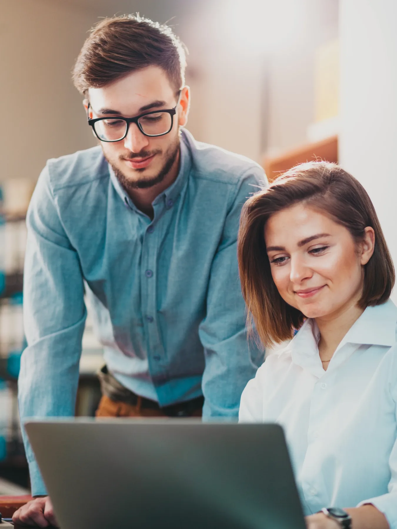Two professionals collaborate over a laptop in a modern office setting, with one person standing and the other seated, both focused on the screen, symbolizing teamwork and research at Alimentiv.