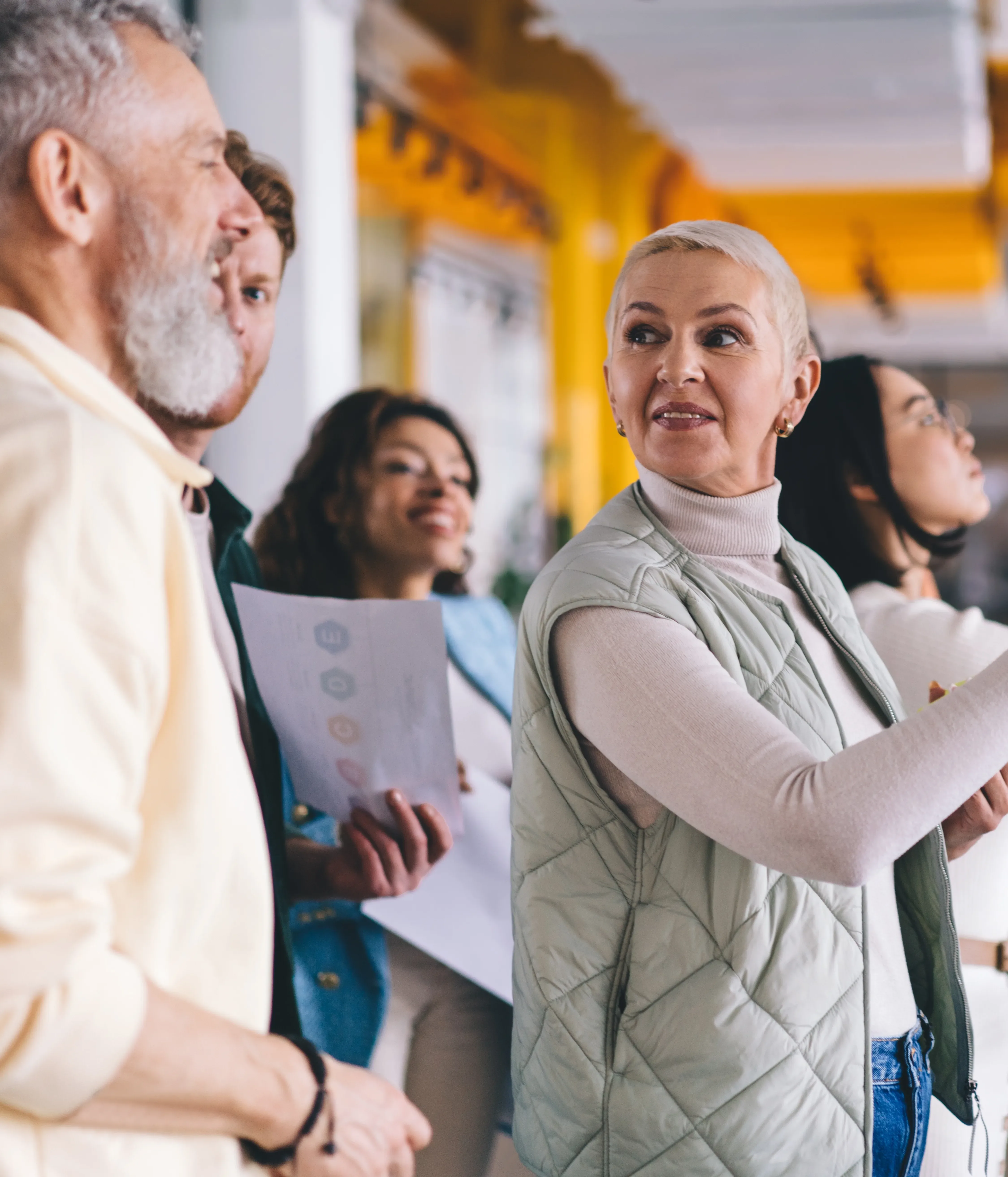 A diverse group of professionals collaborates in a modern workspace, with a senior woman placing notes on a glass wall while colleagues observe and discuss ideas, representing strategic planning and teamwork at Alimentiv
