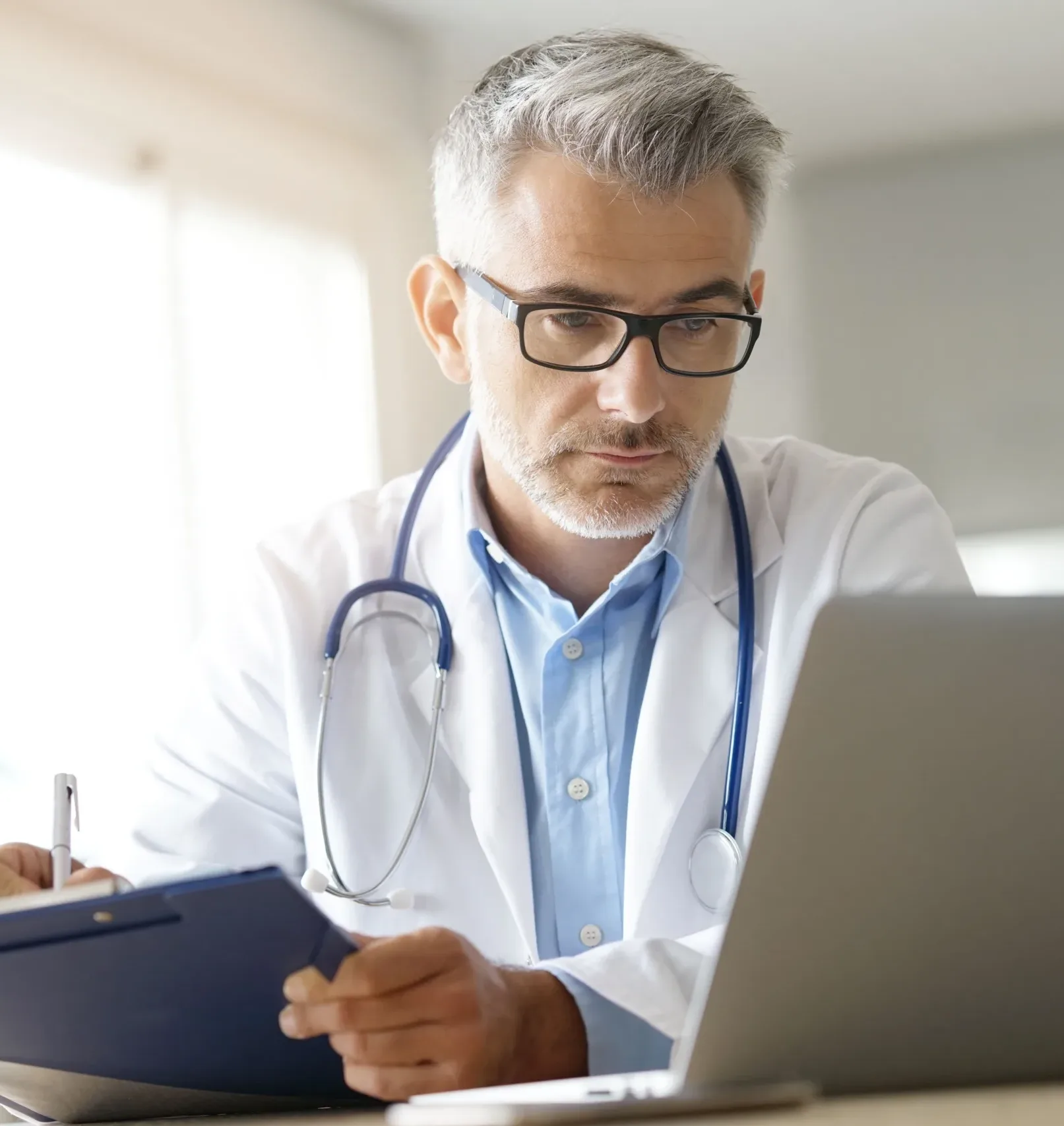 A physician wearing a white coat and stethoscope reviews notes on a clipboard while working on a laptop in a bright clinical office, representing focused medical research and data analysis at Alimentiv